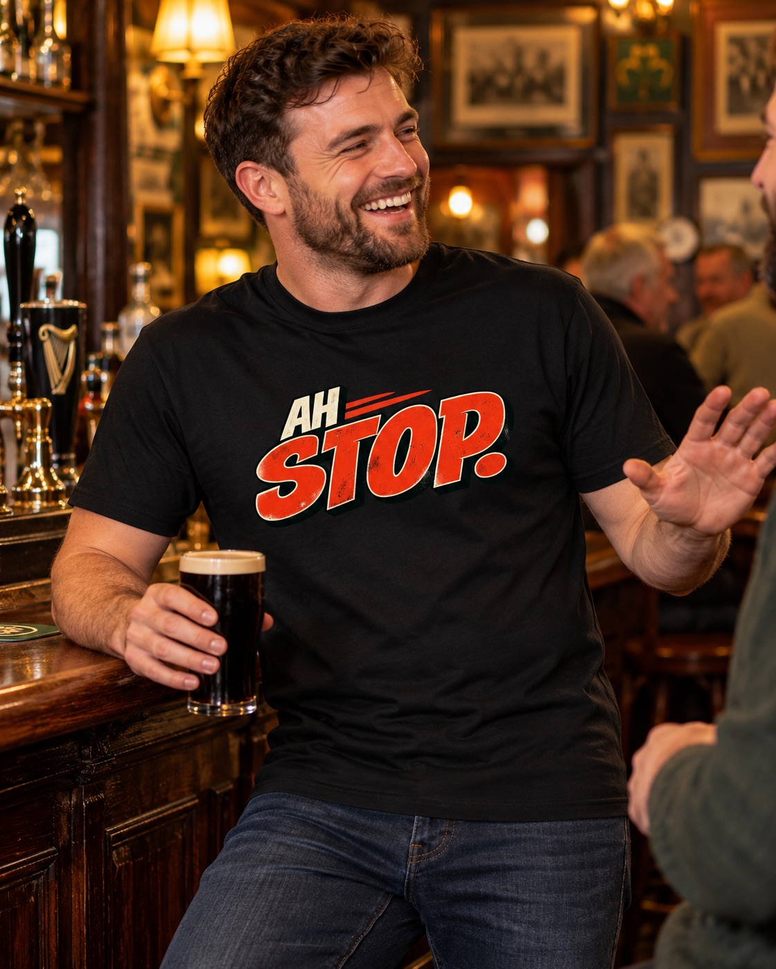 Smiling man wearing a black Ah Stop Irish humour T-shirt while holding a pint and chatting with a friend inside a traditional pub