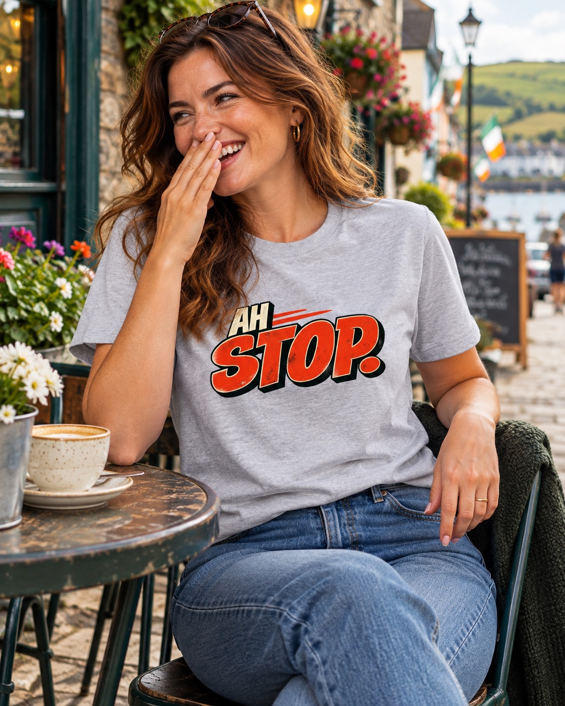 Woman wearing ash grey Ah Stop Irish graphic T-shirt and jeans, laughing at an outdoor café table beside a cup of coffee