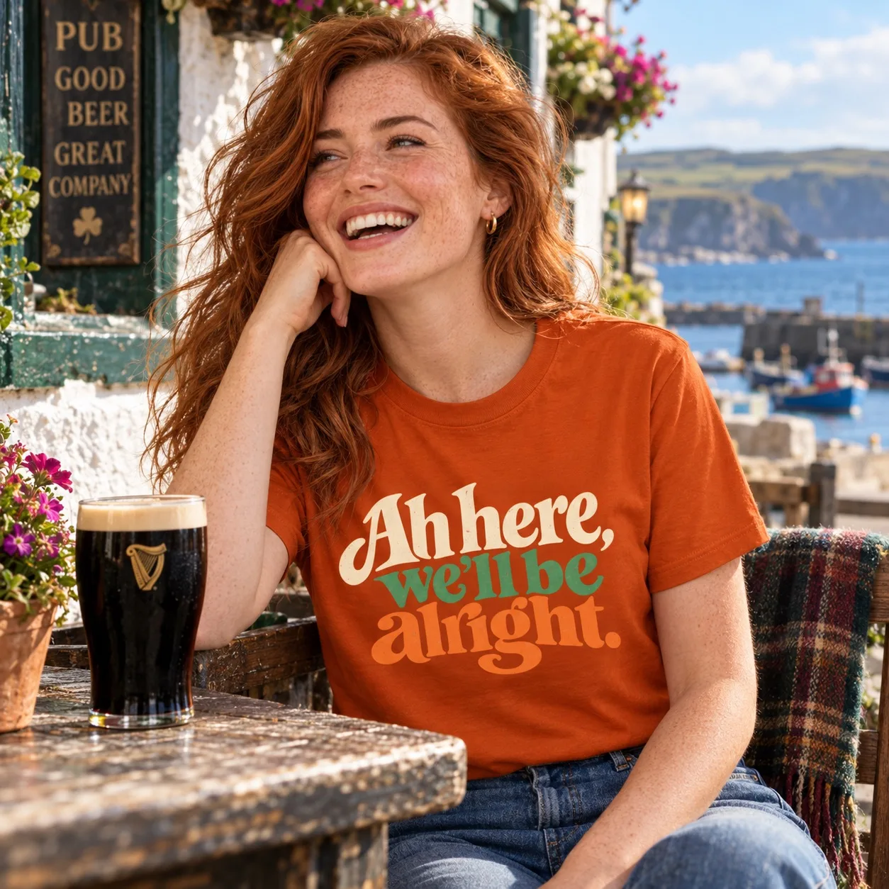 Woman wearing the orange Ah Here, We’ll Be Alright Irish T-Shirt beside an Irish harbour pub