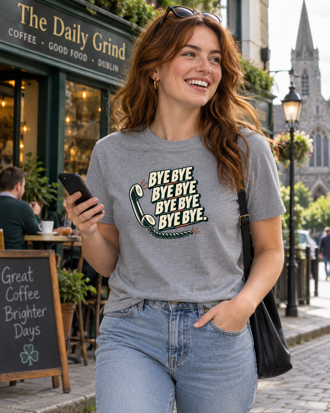 Smiling woman wearing ash grey Bye Bye Bye Bye Irish T-shirt and jeans while holding her mobile phone outside a Dublin café