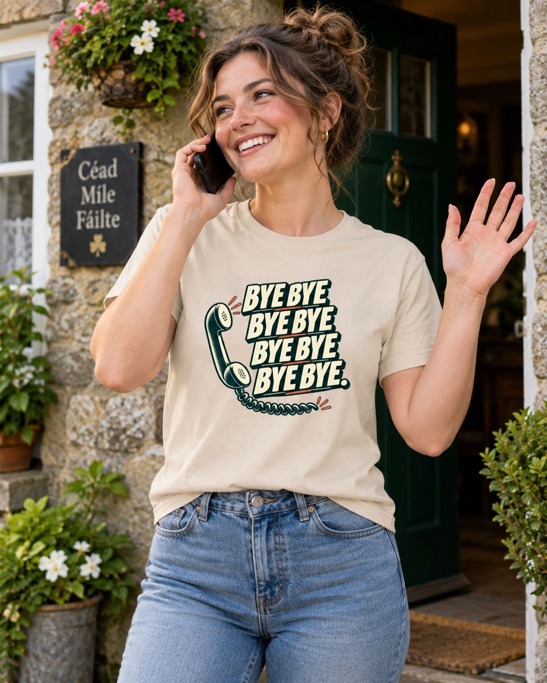 Woman wearing natural cream Bye Bye Bye Bye Irish saying T-shirt, smiling and waving while talking on her phone outside a stone building