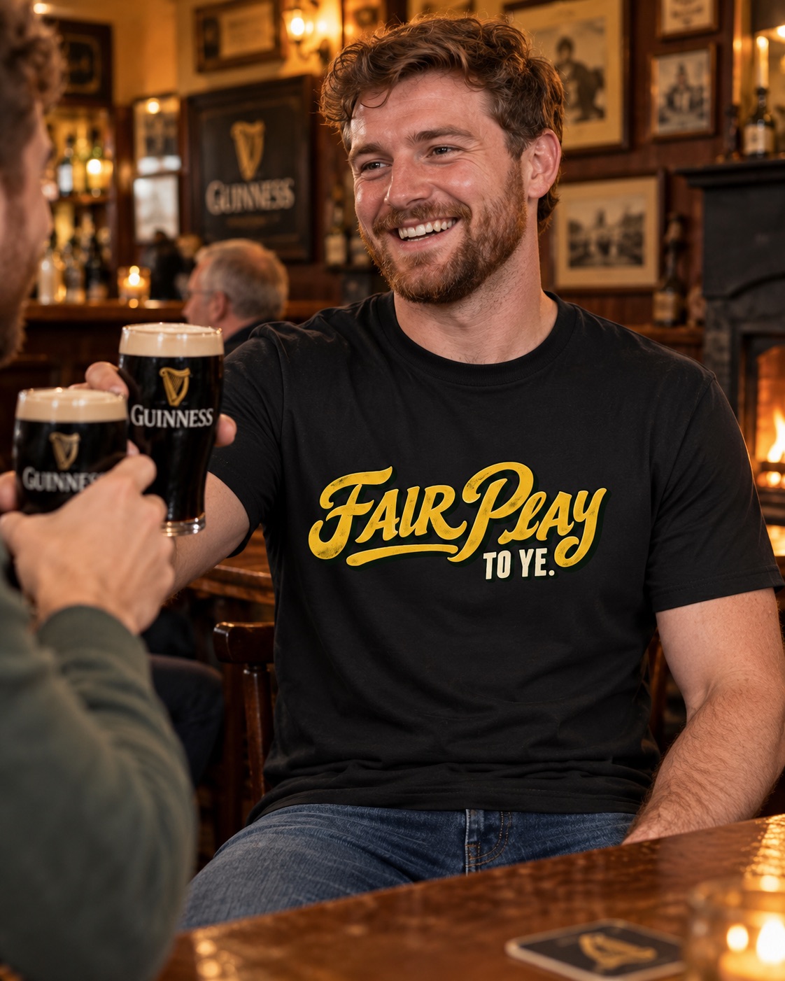 Smiling man wearing black Fair Play To Ye Irish graphic T-shirt while raising a pint of Guinness with a friend in a traditional pub