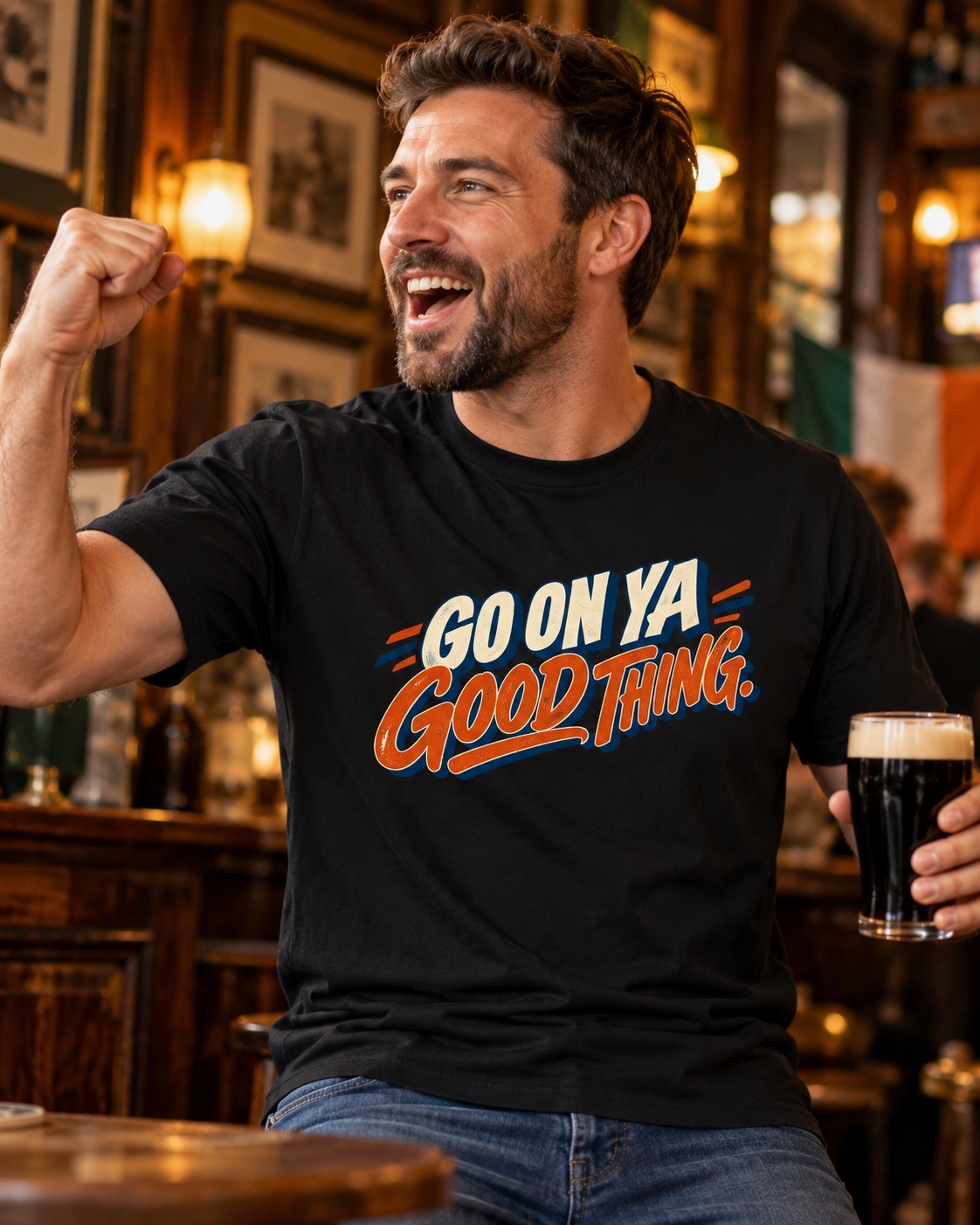 Smiling man wearing black Go On Ya Good Thing Irish T-shirt, raising his fist and holding a pint inside a traditional pub