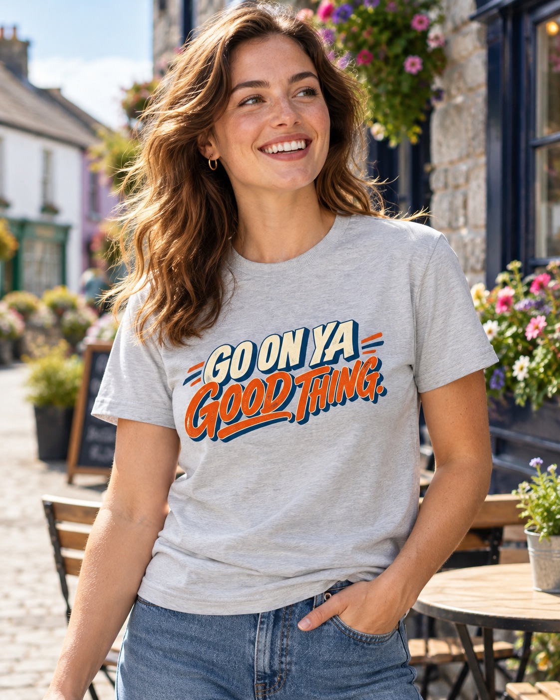 Woman wearing ash grey Go On Ya Good Thing Irish saying T-shirt with jeans outside a village café decorated with flowers