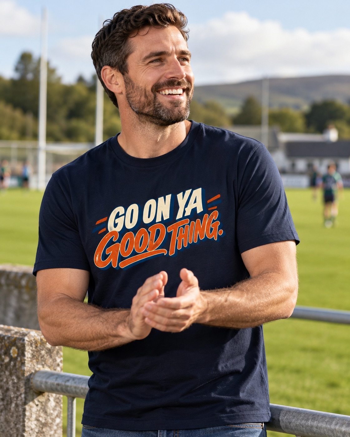 Man wearing navy Go On Ya Good Thing Irish graphic T-shirt, smiling and applauding beside a GAA pitch