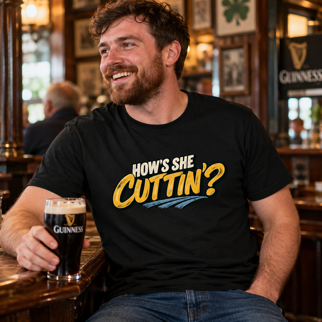 Smiling man wearing black How's She Cuttin' Irish T-shirt while holding a pint of Guinness inside a traditional pub