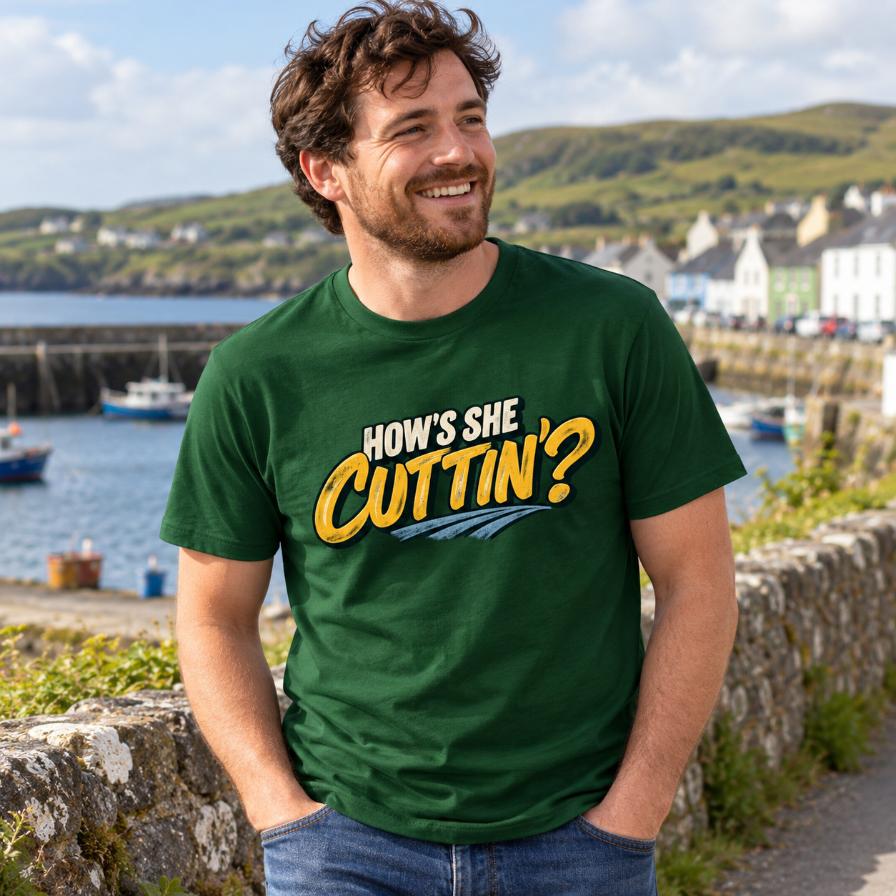 Smiling man wearing forest green How's She Cuttin' Irish graphic T-shirt and jeans beside a coastal harbour