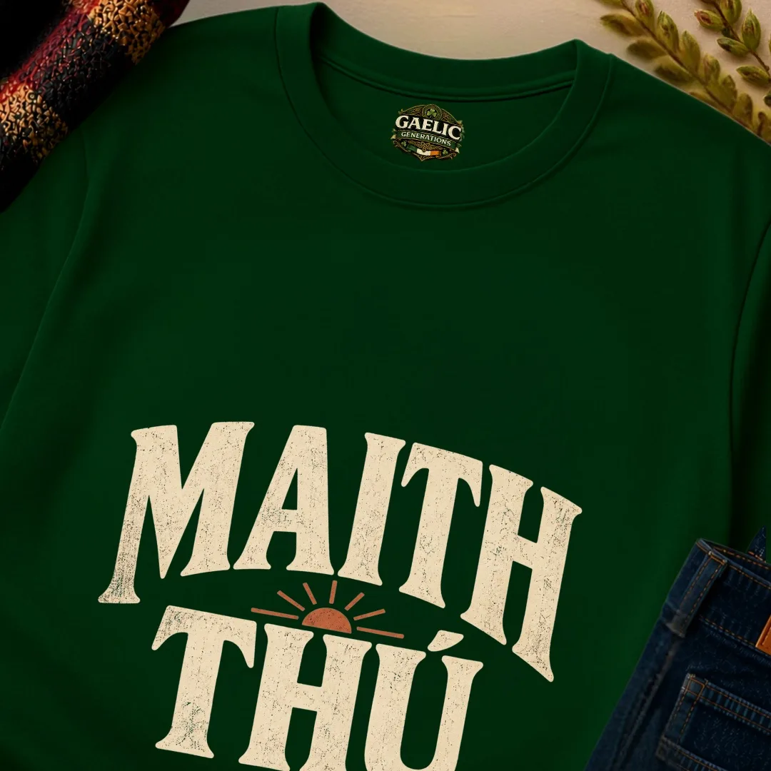 Close-up of forest green Maith Thú Irish T-shirt with cream lettering and rust sunrise graphic