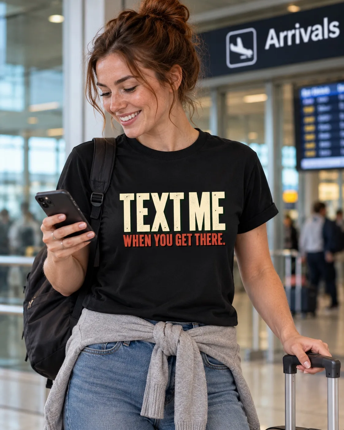 Woman wearing a black Text Me When You Get There T-shirt while checking her phone at airport arrivals